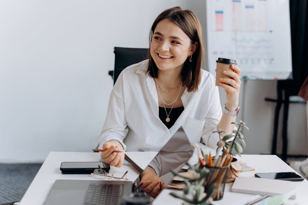 Smiling woman holding coffee cup at modern office desk, exuding happiness and productivity.
