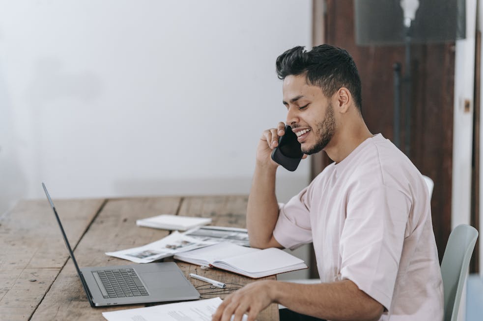 Young Hispanic man engaged in a phone call while working remotely on a laptop at home.