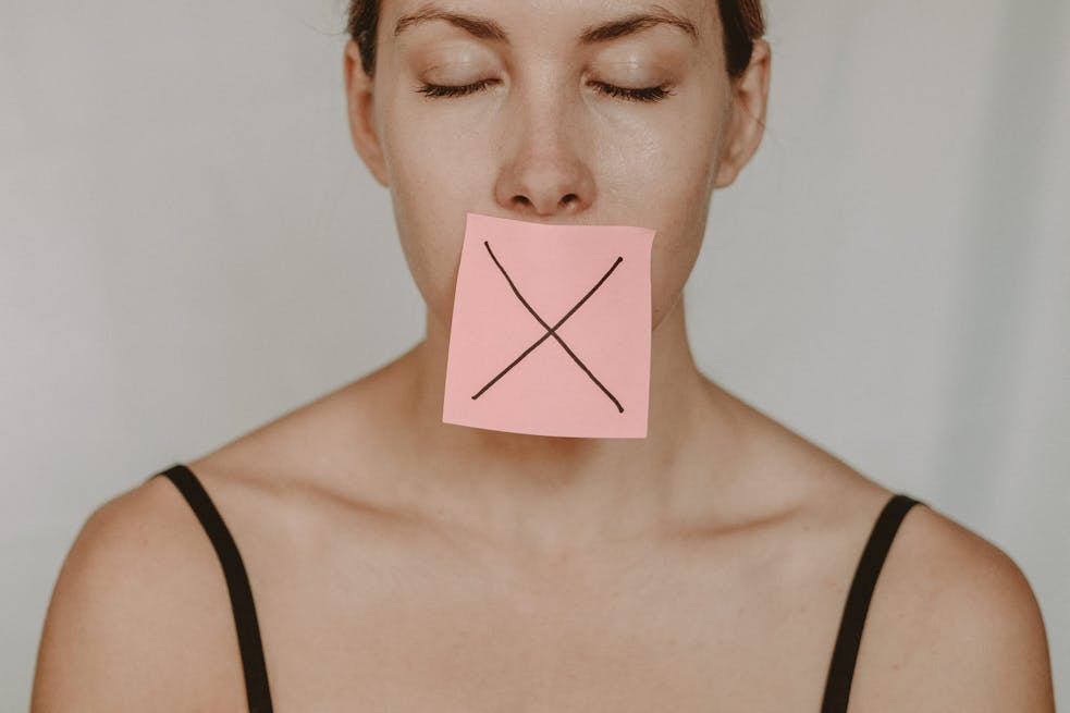 A woman with eyes closed and a note with an X on her mouth, signifying silence or prohibition.