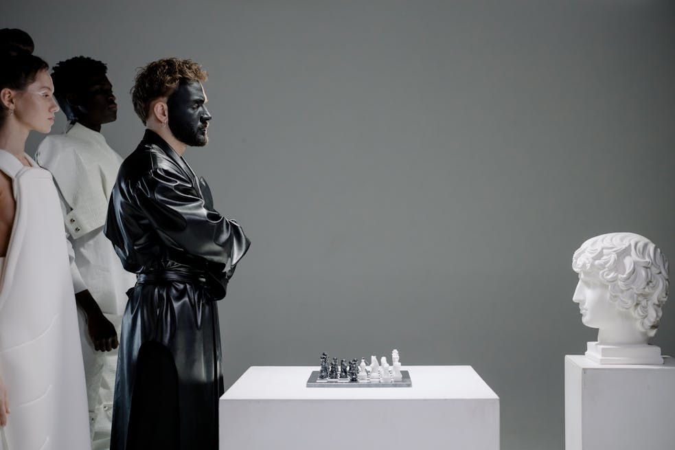 Dramatic studio portrayal of chess game, featuring performers in costume and face paint, alongside a classic sculpture.