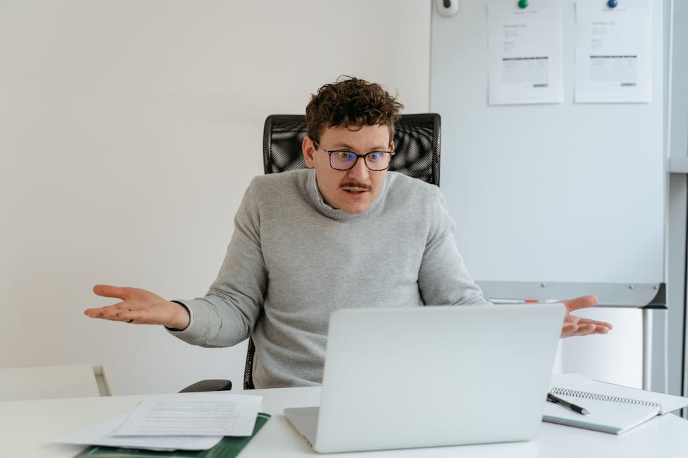 A man in a gray sweater and glasses gesturing at his laptop screen during a video call in an office setting.